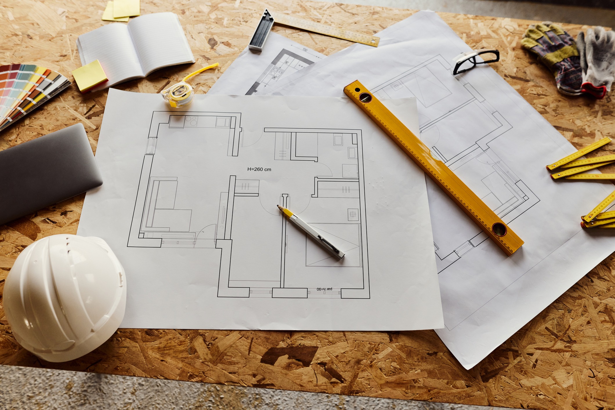 A wooden table with architectural blueprints, a pen, a hard hat, a tape measure, a yellow ruler, gloves, a laptop, a notebook, and color swatches, suggesting a construction or design project in progress.