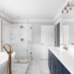 Modern bathroom with a freestanding tub, glass-enclosed shower, white marble surfaces, gold fixtures, navy blue vanity cabinets, and large mirrors. Natural light enters through a window with white blinds.