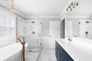 Modern bathroom with a freestanding tub, glass-enclosed shower, white marble surfaces, gold fixtures, navy blue vanity cabinets, and large mirrors. Natural light enters through a window with white blinds.