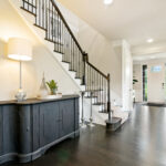Bright entryway of a home with dark hardwood floors, a staircase with black railings, a dark console table with decor and lamp, and a white front door letting in natural light.