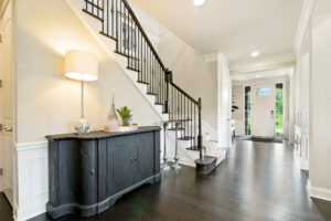 Bright entryway of a home with dark hardwood floors, a staircase with black railings, a dark console table with decor and lamp, and a white front door letting in natural light.