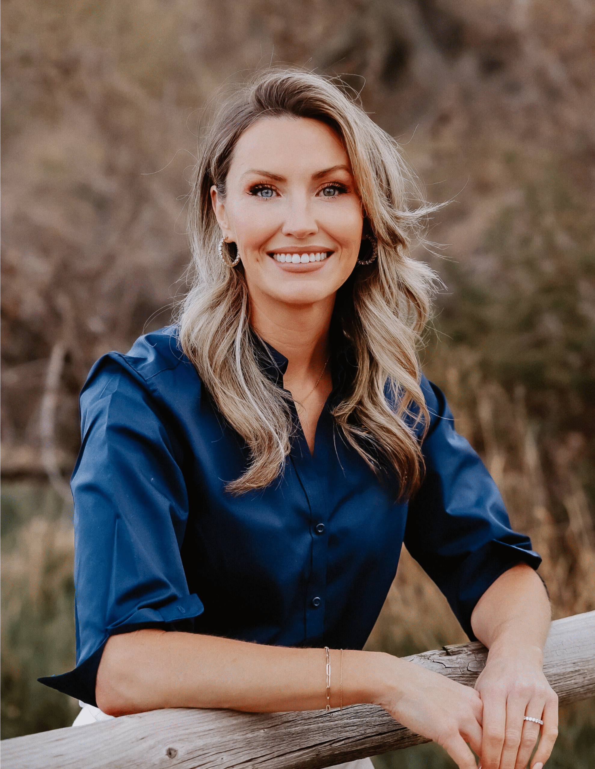 A woman with long blonde hair, wearing a navy blue blouse, smiles while leaning on a wooden fence outdoors with trees and foliage in the background.
