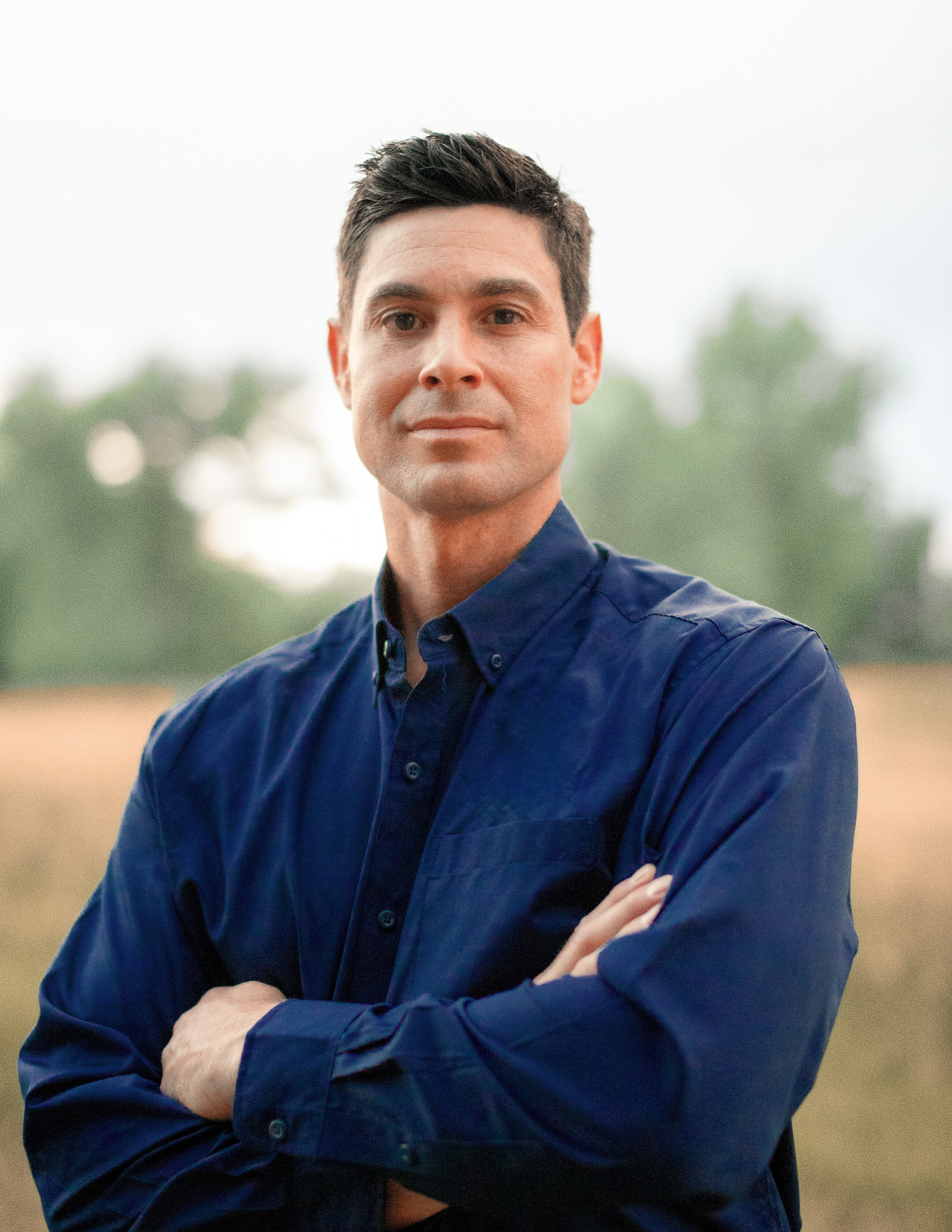 A man with short dark hair, wearing a dark blue button-up shirt, stands outdoors with his arms crossed. The background is blurred with green trees and a field.