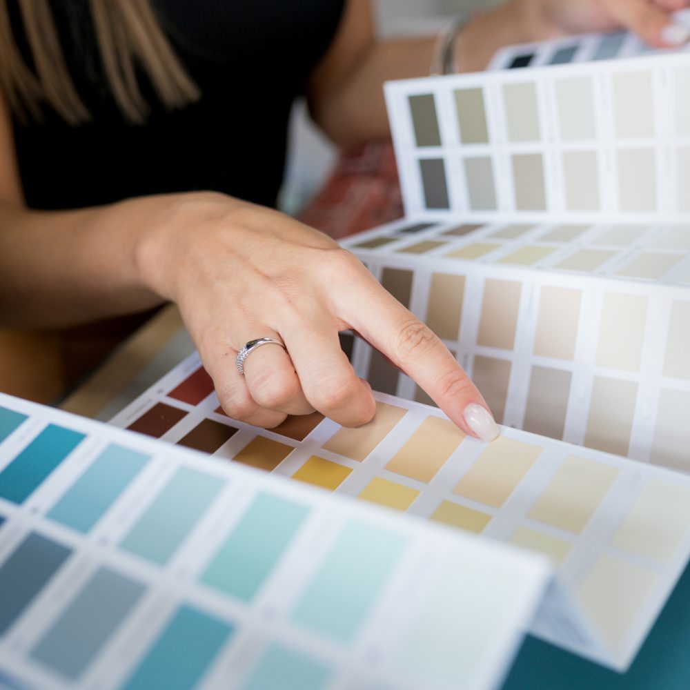 A person points to a color swatch on a paint sample chart, surrounded by various color options, while another hand holds part of the chart.