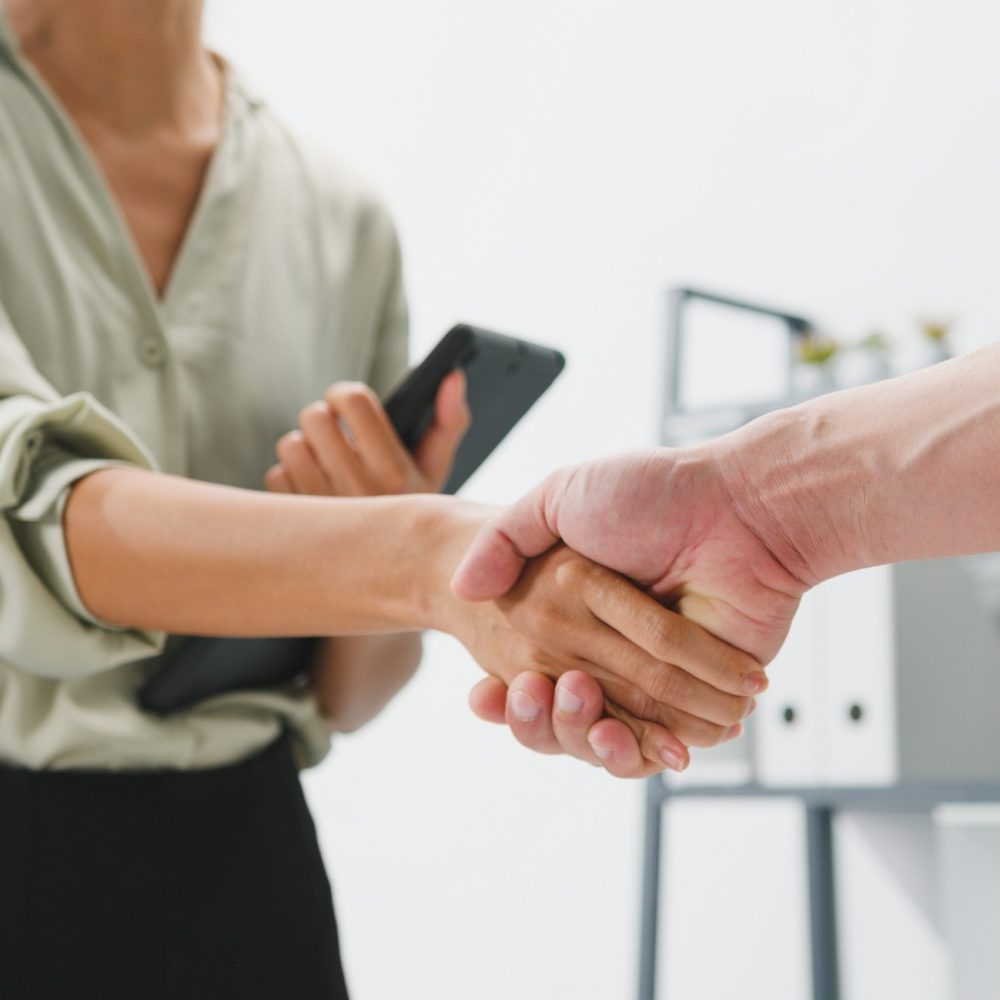 Two people shaking hands in an office setting; one person holds a tablet, and only their upper bodies and hands are visible. The background shows shelves with binders and plants.