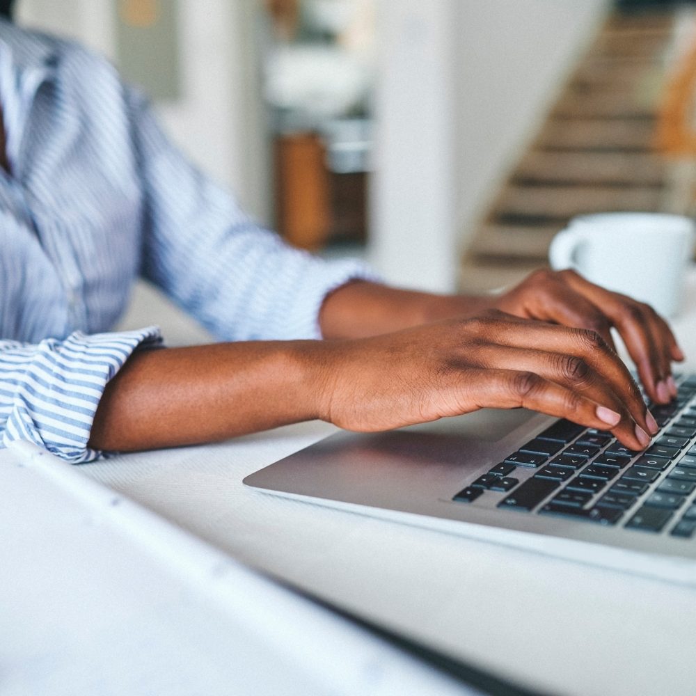 A person wearing a blue and white striped shirt types on a laptop at a table, with a white coffee mug and notebook nearby. The background shows a staircase and a softly lit interior.