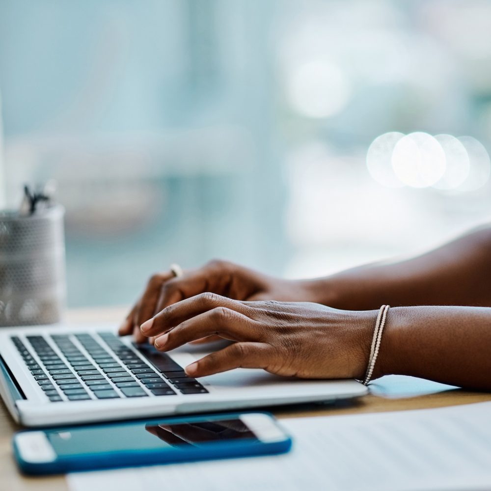 A person types on a laptop at a desk with a smartphone, papers, and a pen holder nearby. The scene is well-lit with natural light from a window in the background.