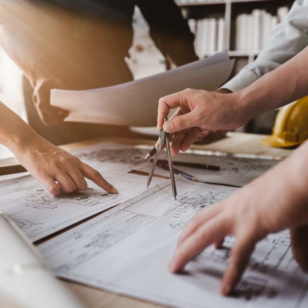 Close-up of people’s hands pointing to and discussing architectural blueprints on a table, with drafting tools and a yellow safety helmet visible, suggesting a collaborative planning or design meeting.