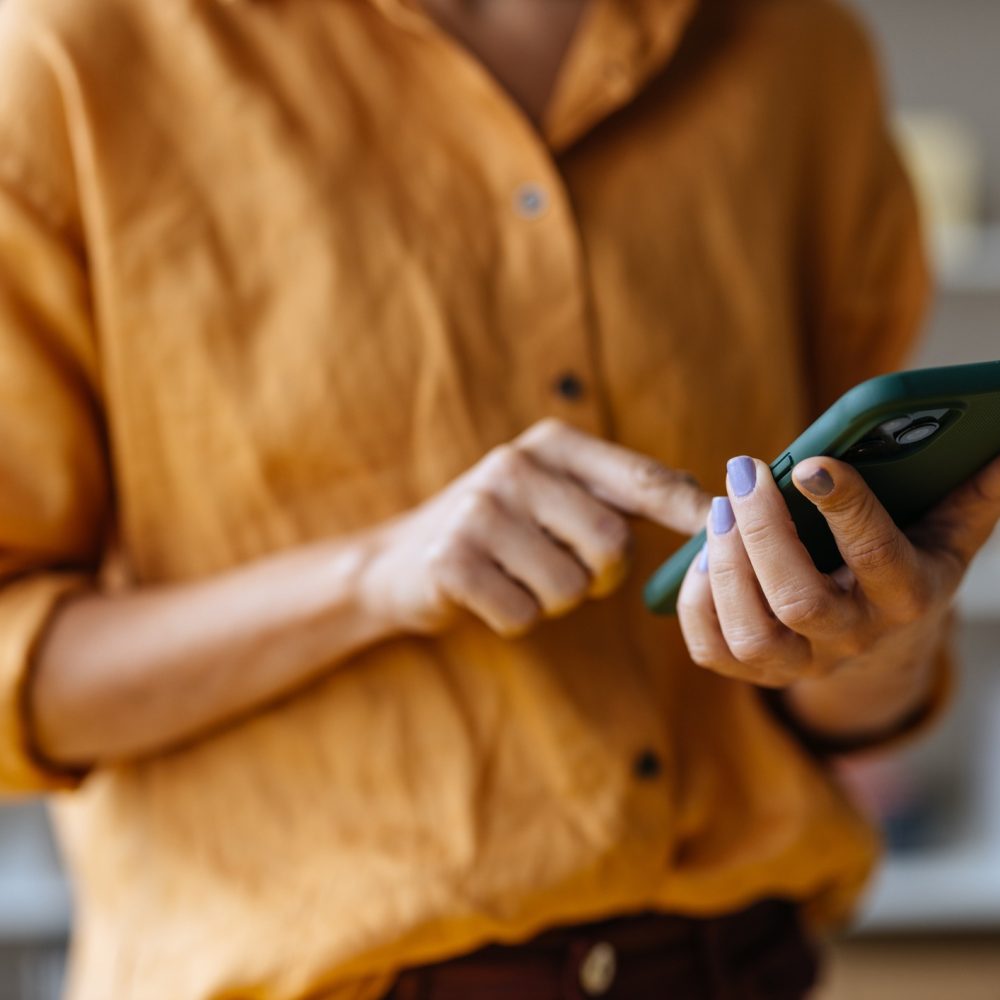 A person wearing a mustard yellow shirt uses a smartphone. The focus is on their hands and phone, with their face out of frame. The person has light purple nail polish. The background is softly blurred.