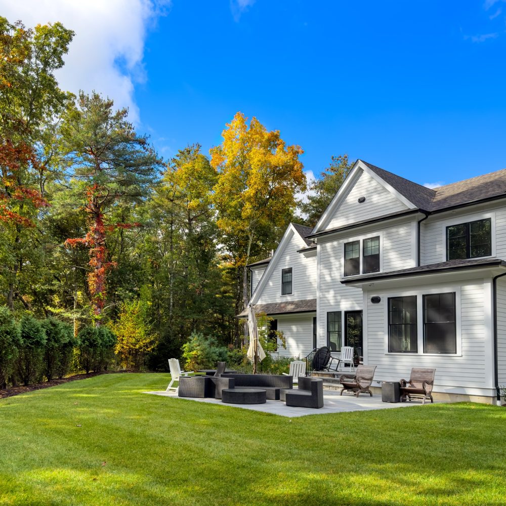 A modern white two-story house with large windows sits next to a green lawn and patio area with outdoor seating, surrounded by trees with early autumn foliage under a blue sky.