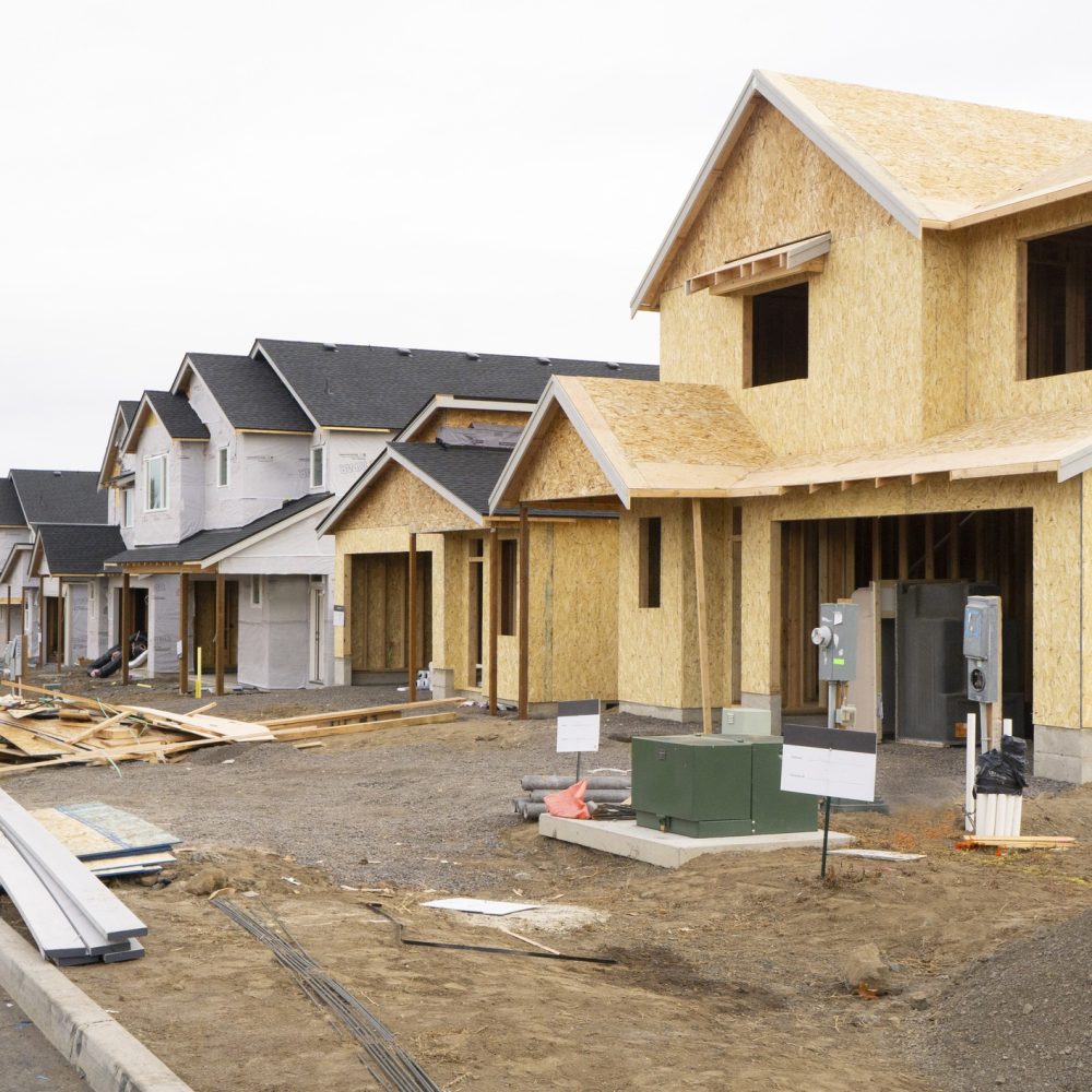 A row of new houses under construction in a suburban neighborhood, with plywood exteriors, construction materials, and equipment scattered around the unfinished driveways.
