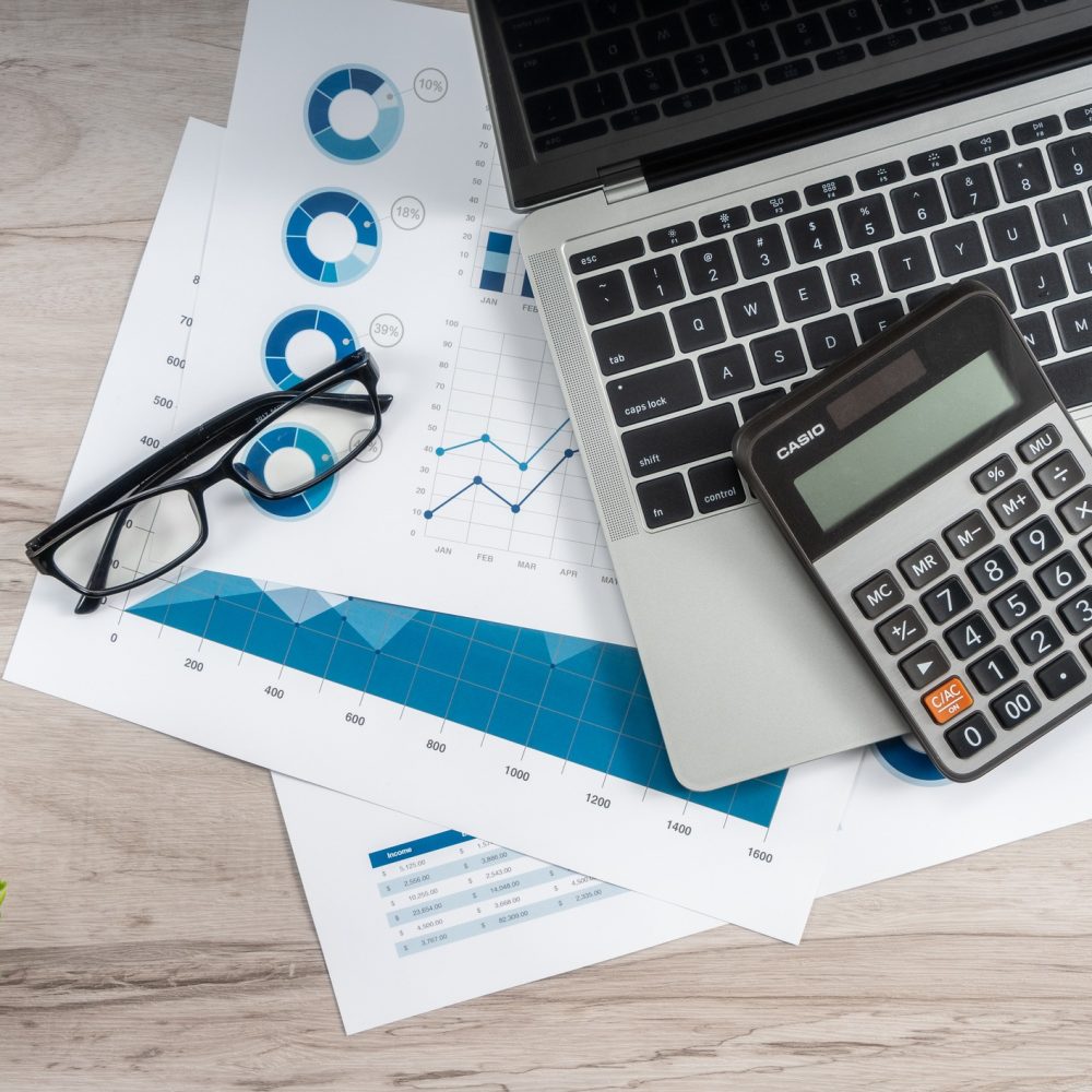 A laptop, calculator, and eyeglasses rest on printed financial charts and graphs on a wooden desk, with a small potted plant nearby.