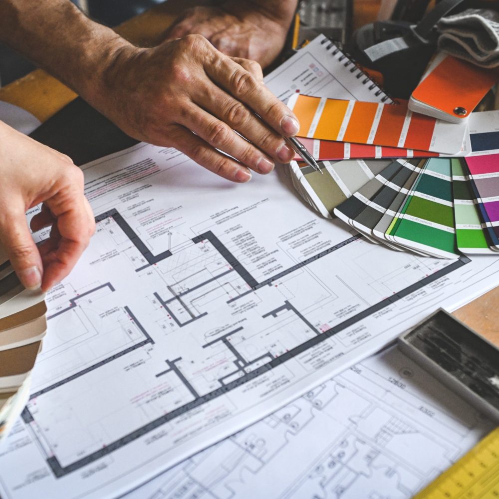 Two people review a building floor plan while selecting paint colors from multiple color swatches on a cluttered wooden desk, suggesting an interior design or renovation planning process.