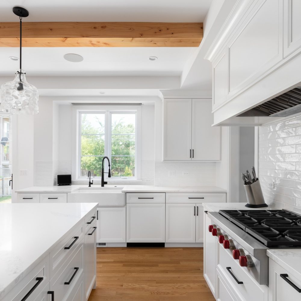 Modern white kitchen with large island, marble countertops, gas stove, pendant lights, white cabinets, wood floor, and a window letting in natural light. Stainless steel appliances and a black faucet are visible.